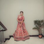 Woman in a vibrant bridal lehenga poses gracefully indoors beside a plant.