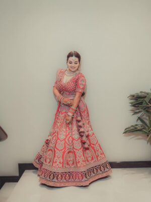 Woman in a vibrant bridal lehenga poses gracefully indoors beside a plant.