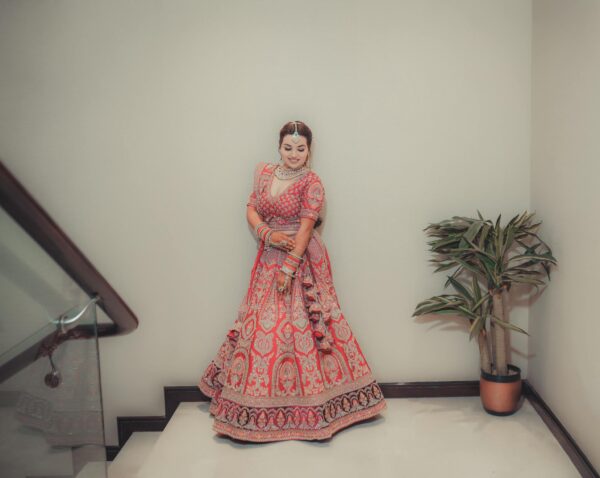 Woman in a vibrant bridal lehenga poses gracefully indoors beside a plant.